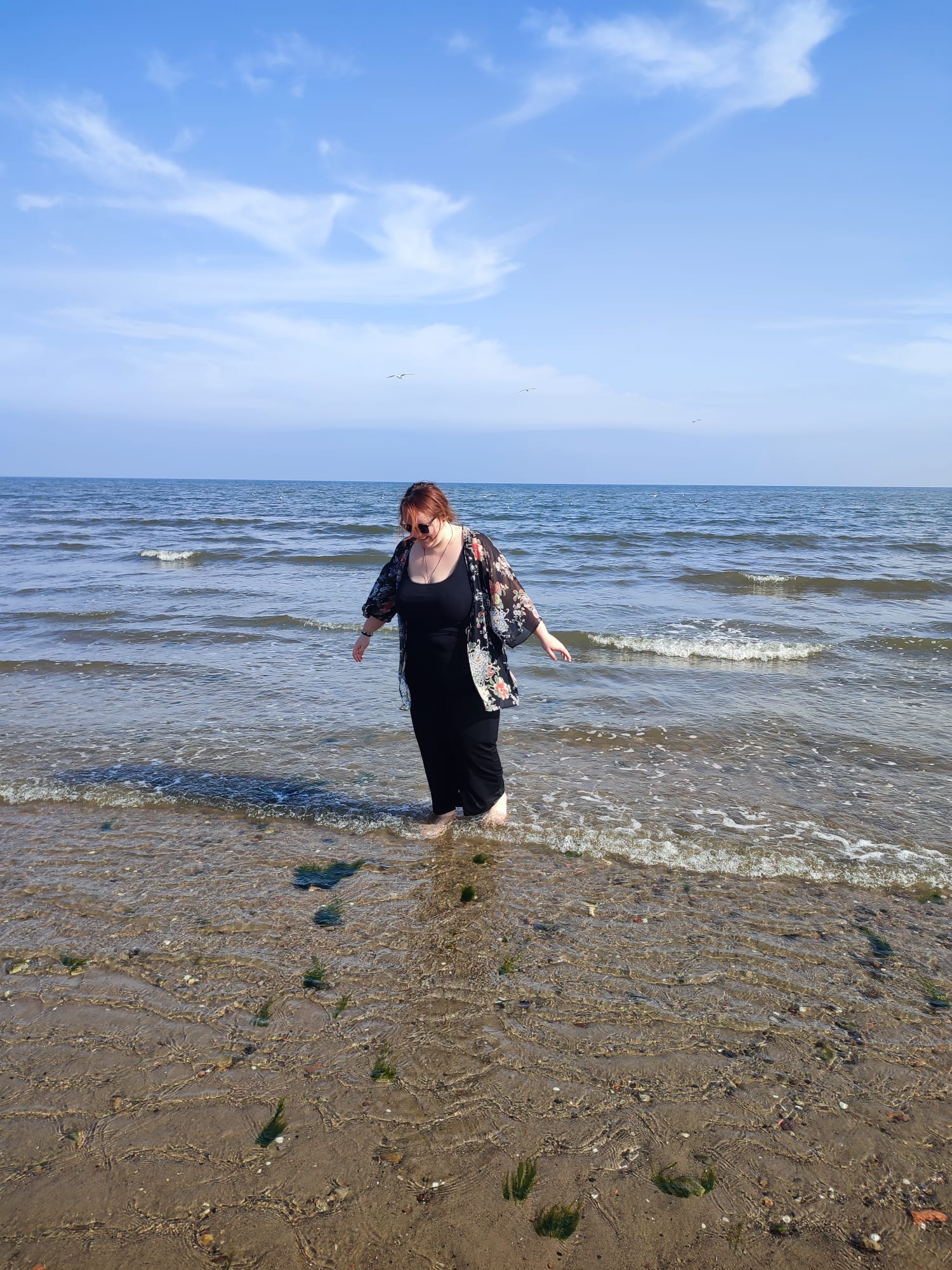Babsi in luftiger Kleidung läuft barfuß am Strand. Hinter ihr sind blauer Himmel und Wellen.